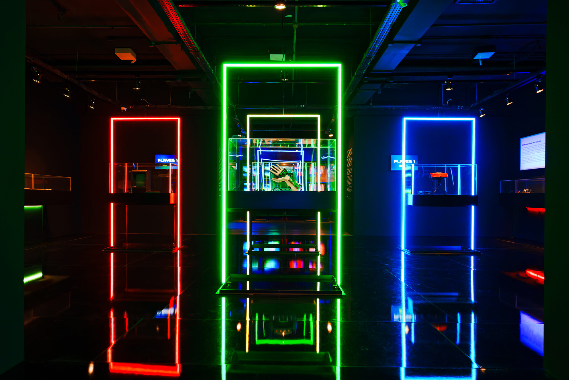 Player 1: 50 Anos dos Videogames · Três pórticos de neon e as vitrines do acervo, com reflexo no piso